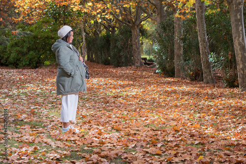 mujer embarazada con gorra blanca, abrigo gris y sosteniendo la ropa de su futuro bebé se encuentra en un parque con colores otoñales. © Ricardo