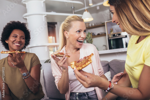 Happy young female friends with wine and pizza on sofa at home.