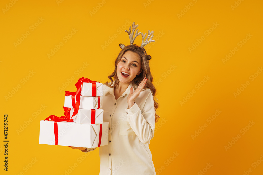 beautiful attractive surprised girl holding in her hands a large pyramid of gift boxes with red ribbons, wearing antlers on her head on an isolated yellow background beautiful attractive surprised girl holding in her hands a large pyramid of gift boxes with red ribbons, wearing antlers on her head on an isolated yellow background