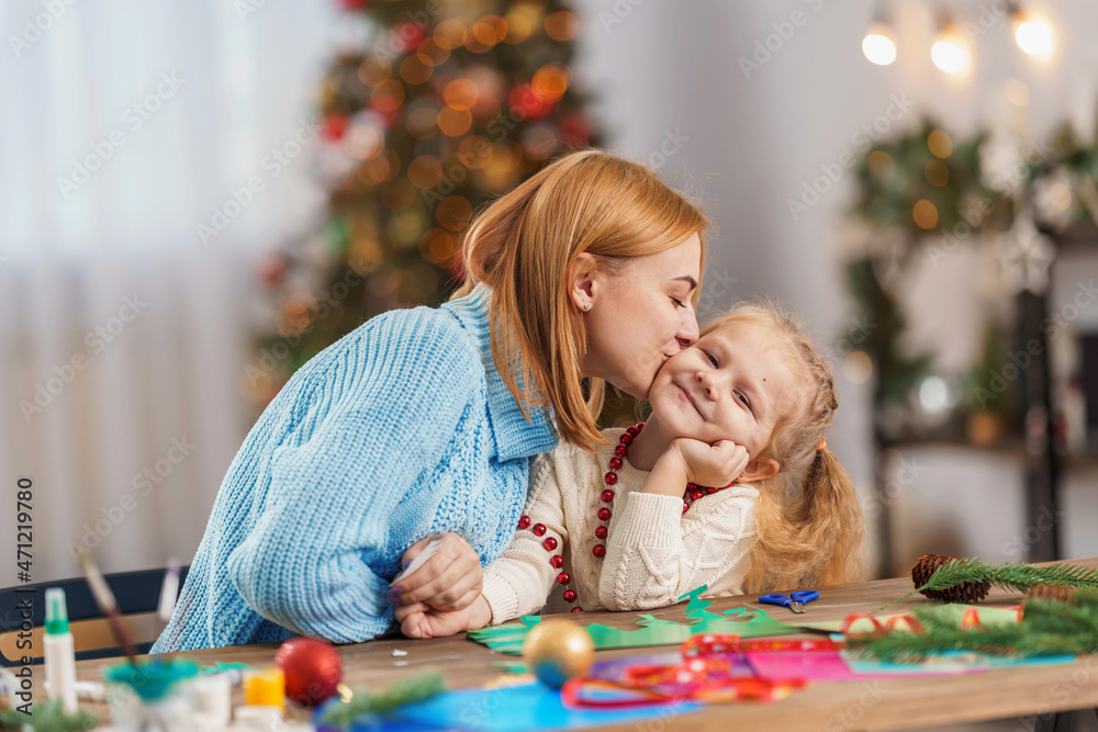 cute little girl and her mother are engaged in needlework, creativity, enjoying the process, sitting at a desk, a lot of materials, preparing for a holiday in a decorated house.