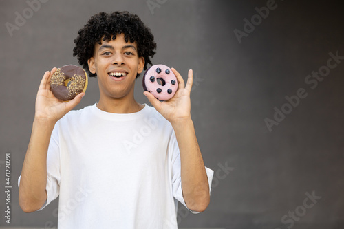 Dessert, fast food, fun and people concept - Smiling African-American guy holding a chocolate and blueberry doughnut near his face, wearing a white T-shirt on a gray background with copy space