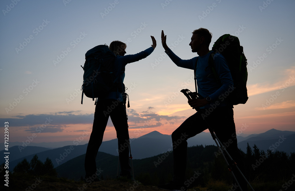 Family pair of hikers fun clapping each other's hands when they reached top of the mountain during outdoor hike. Evening hiking on mountain hills in twilight under evening sky. Concept of travelling.