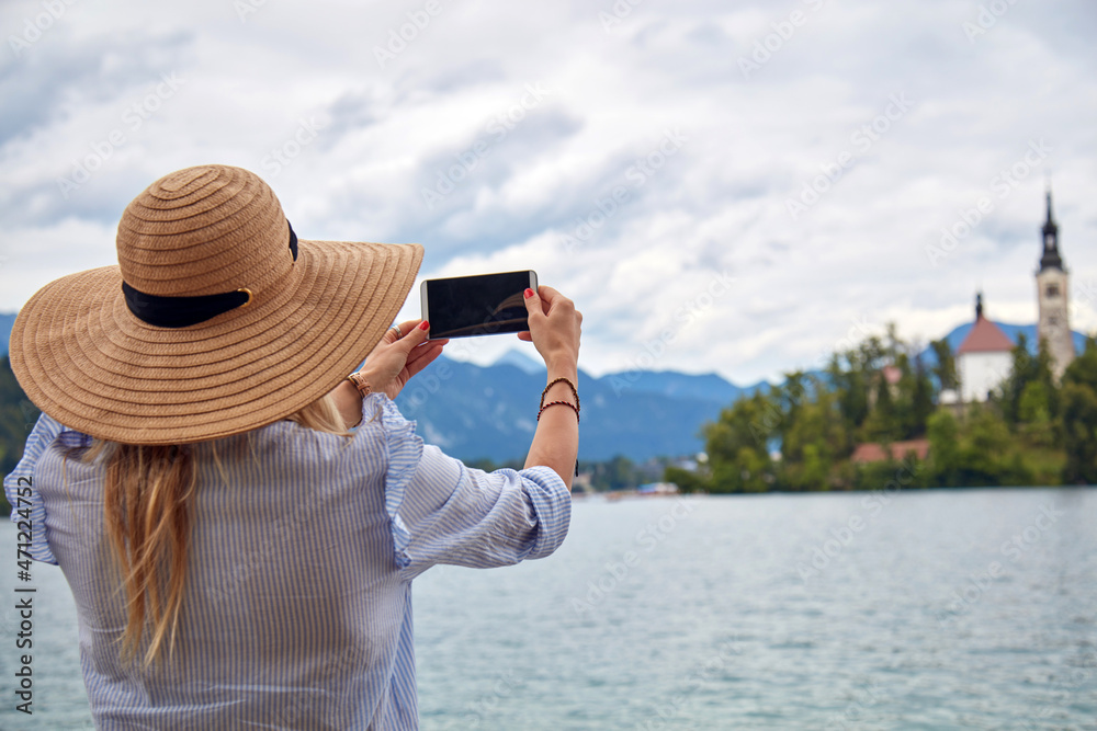 Fototapeta premium Young adult tourist woman enjoying on a lake while taking photos with a modern smartphone.