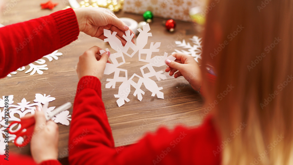 child with his mother carves snowflake for Christmas. family makes decorations for new year. Children's hands carve snowflake on background wooden table. Christmas theme. Selective focus. Rear view child with his mother carves snowflake for Christmas. family makes decorations for new year. Children's hands carve snowflake on background wooden table. Christmas theme. Selective focus. Rear view