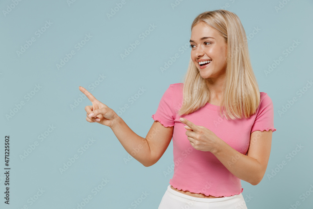Young smiling blonde woman 20s wearing casual pink t-shirt point index finger aside on workspace area mock up isolated on plain pastel light blue background studio portrait. People lifestyle concept. Young smiling blonde woman 20s wearing casual pink t-shirt point index finger aside on workspace area mock up isolated on plain pastel light blue background studio portrait. People lifestyle concept.