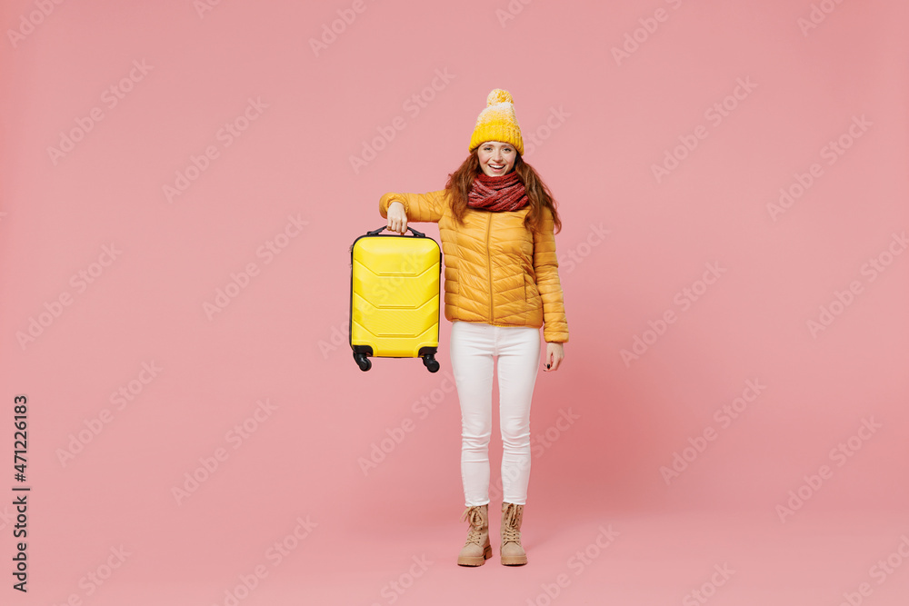 Full size body length swanky cheerful young woman 20s years old wears yellow jacket hat mittens looking camera hold showing suitcase bag isolated on plain pastel light pink background studio portrait.