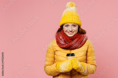 Smiling young woman 20s years old wears yellow jacket hat mittens look camera hold takeaway delivery craft paper brown cup coffee to go isolated on plain pastel light pink background studio portrait.