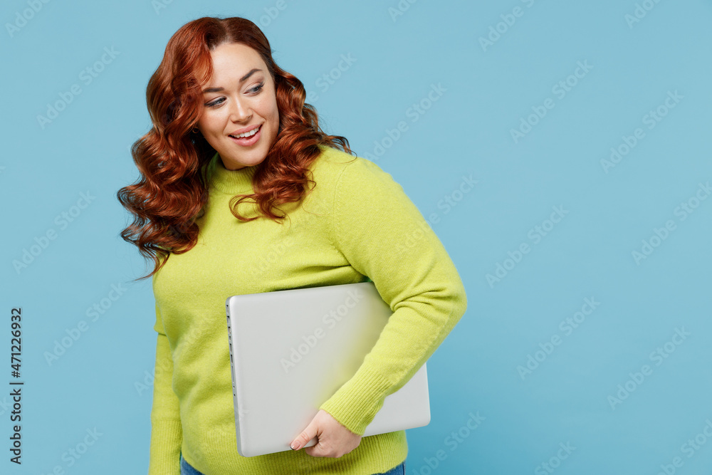 Obraz premium Young side view chubby overweight plus size big fat fit woman wear green sweater hold closed laptop pc computer look aside isolated on plain blue background studio portrait. People lifestyle concept.