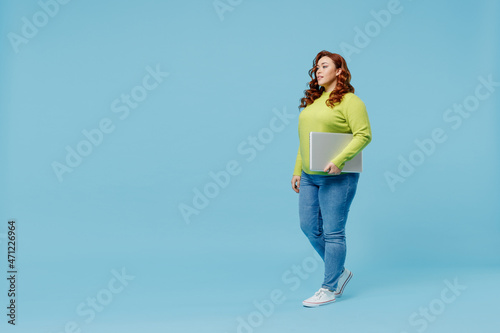 Full body side view young chubby overweight plus size big fat fit woman 20s wear green sweater hold closed laptop pc computer walking going isolated on plain blue background. People lifestyle concept.
