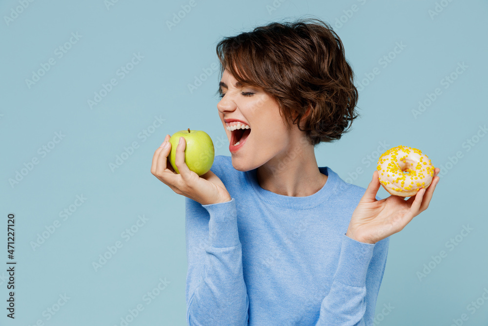 Φωτογραφία Young woman wearing casual sweater look camera hold donut dessert bite apple choose beatween healthy and junk food isolated on plain pastel light blue background studio