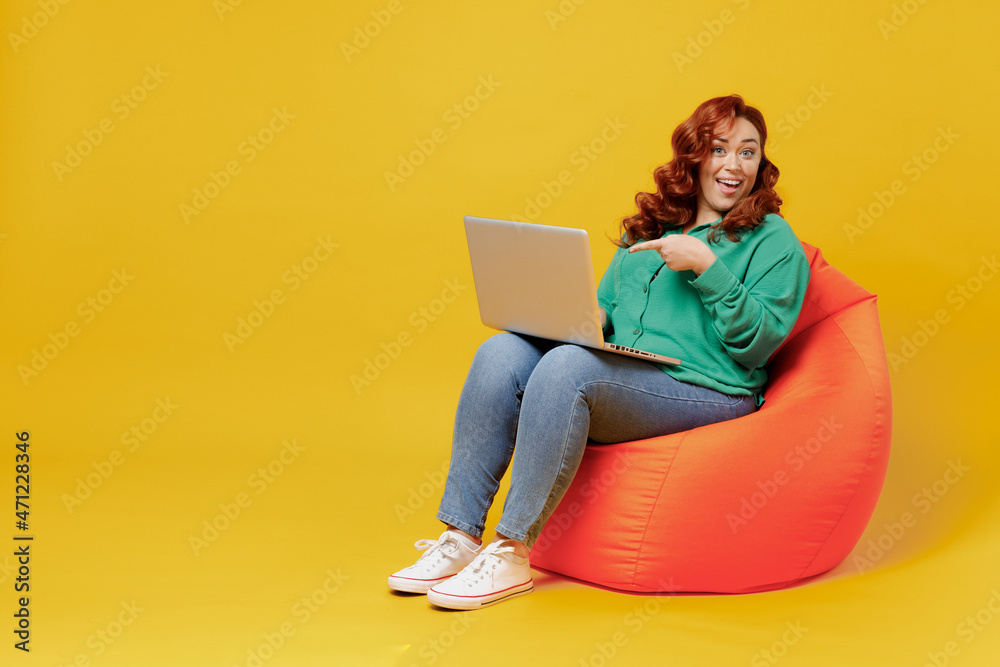 Full size body length young ginger chubby overweight woman 20s wears green shirt sit in bag chair hold use work point finger on laptop pc computer isolated on plain yellow background studio portrait. Full size body length young ginger chubby overweight woman 20s wears green shirt sit in bag chair hold use work point finger on laptop pc computer isolated on plain yellow background studio portrait.