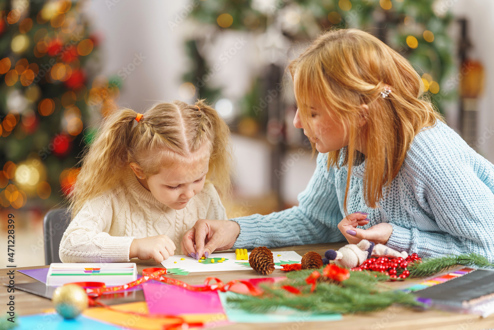 cute little girl and her mother are engaged in needlework, creativity, enjoying the process, sitting at a desk, a lot of materials, preparing for a holiday in a decorated house.