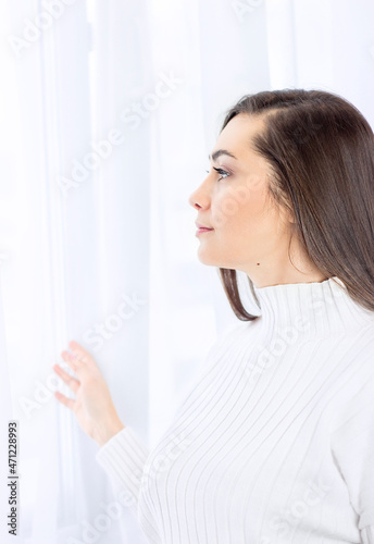 a girl in white stands and looks out the window