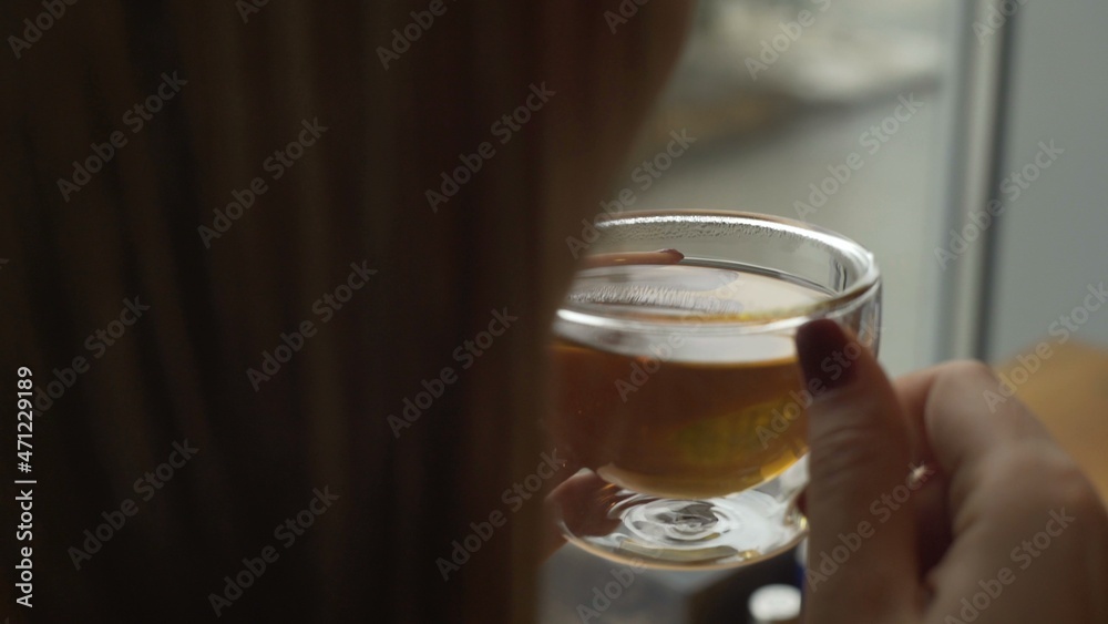 A young girl is sitting near the window and holding a glass mug with warm tea blowing cool air into her cup A young girl is sitting near the window and holding a glass mug with warm tea blowing cool air into her cup