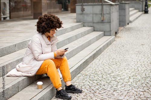 Portrait of Young, cute caucasian woman 40 years old, Sport casual clothes, Sitting on steps, Disposable Cup of coffee. Afro Hairstyle. texting massage using smart phone. photo