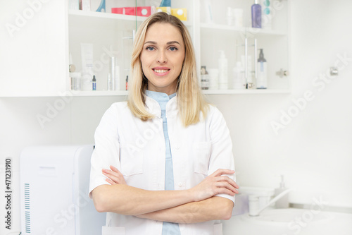 Portrait of a female doctor standing in her office in a clinic. A blonde woman beautician stands with her arms crossed and looks at camera with a smile gainst the background of the medical office.