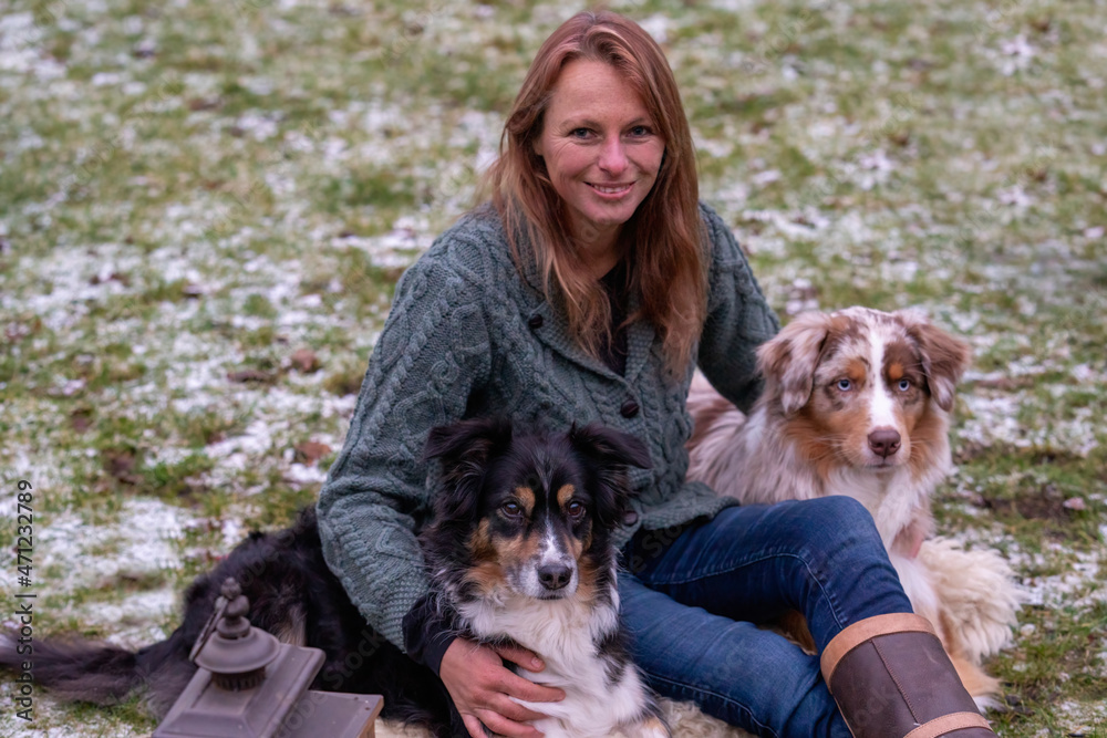 Young woman is sitting outside in the woods with her two Australian Shepherd dogs. Snow on the grass, twilight by the campfire. Cuddles and companion animals Young woman is sitting outside in the woods with her two Australian Shepherd dogs. Snow on the grass, twilight by the campfire. Cuddles and companion animals