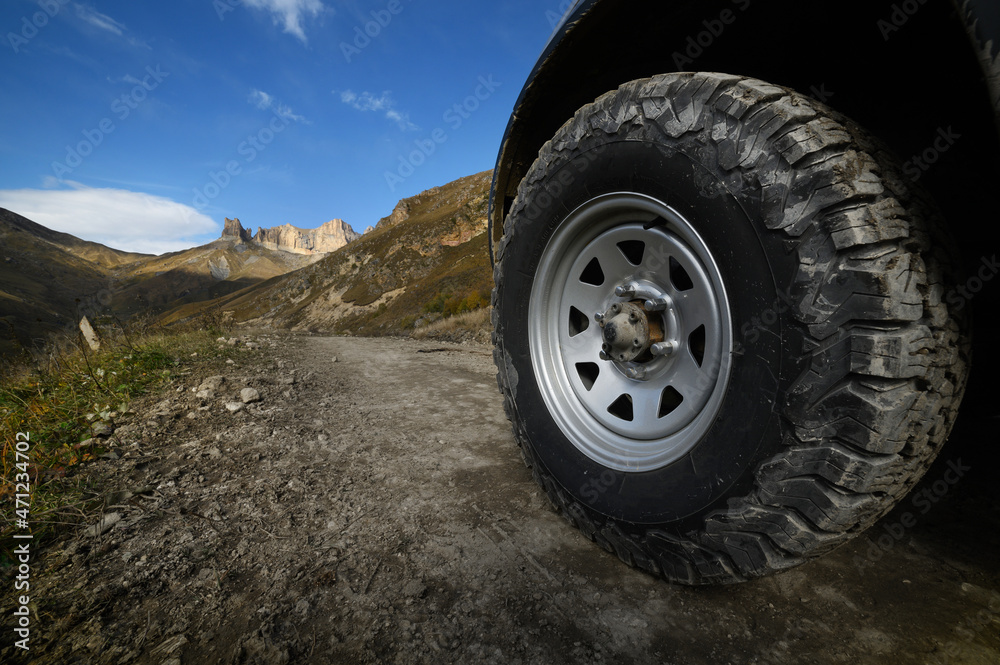 Close-up A large off-road all-terrain car wheel stands on a rough road in the mountains against the backdrop of rocks. Off-road tours and off-road travel concept Close-up A large off-road all-terrain car wheel stands on a rough road in the mountains against the backdrop of rocks. Off-road tours and off-road travel concept