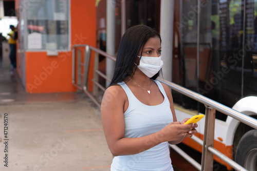 Young woman holding her phone while waiting for her bus at the station. She is wearing a mask due to the COVID-19 pandemic.