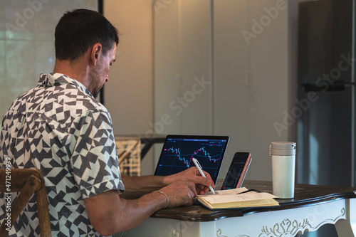 A man trader sits at a table with a laptop and smartphone,  analyzes exchange chart on the screen and writes data to the diary