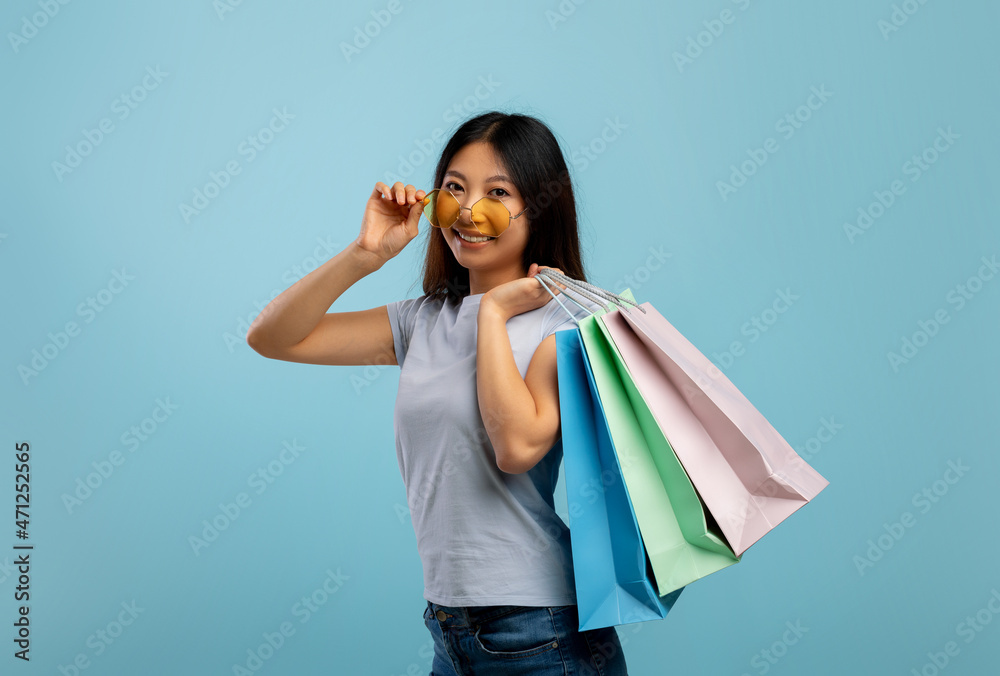 Special offer. Young asian lady touching sunglasses holding colorful shopping bags with purchases, blue background