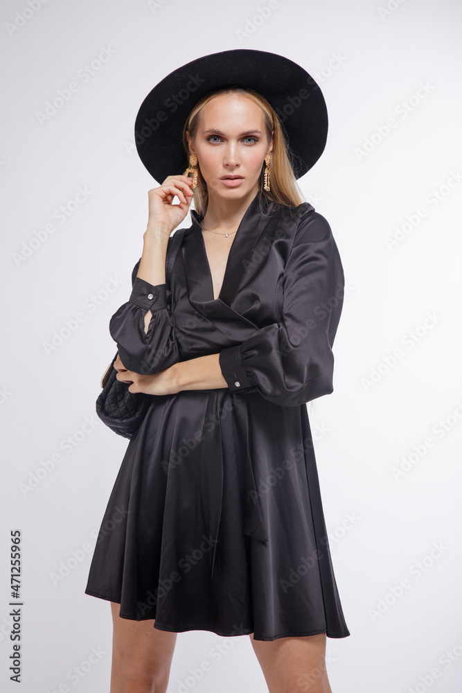 High fashion photo of a beautiful elegant young woman in a pretty black dress, hat, high heels, bag posing over white, soft gray background. Studio Shot, portrait. Blonde, slim figure High fashion photo of a beautiful elegant young woman in a pretty black dress, hat, high heels, bag posing over white, soft gray background. Studio Shot, portrait. Blonde, slim figure