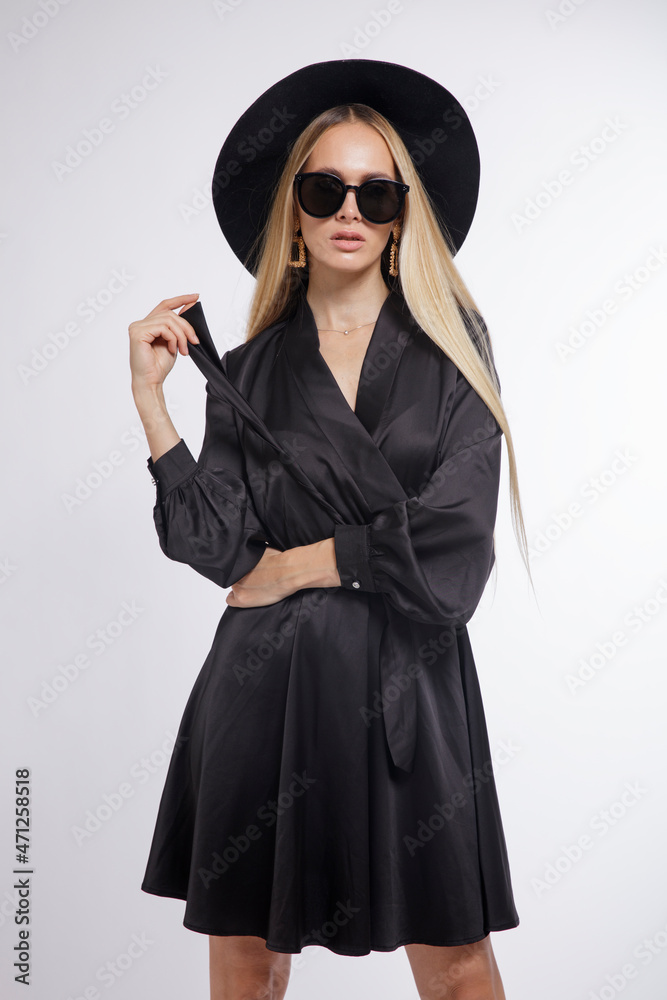 High fashion photo of a beautiful elegant young woman in a pretty black dress, hat, high heels, sunglasses, posing over white, soft gray background. Studio Shot, portrait. Blonde, slim figure