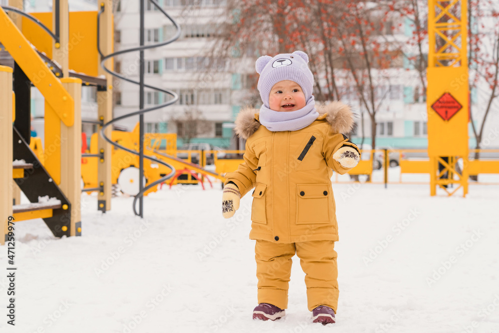 A baby aged 12-17 months on the playground in winter. A kid in a yellow jacket smiles sweetly standing in the snow in the courtyard of an apartment building in Russia. A baby aged 12-17 months on the playground in winter. A kid in a yellow jacket smiles sweetly standing in the snow in the courtyard of an apartment building in Russia.