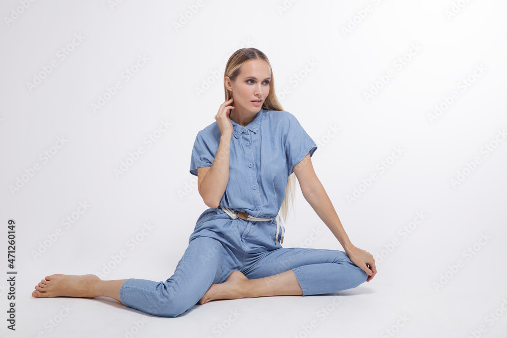 High fashion photo of a beautiful elegant young woman in a pretty blue jumpsuit, barefoot posing over white, soft gray background. Studio Shot, portrait. The model is sitting in the background. Blonde
