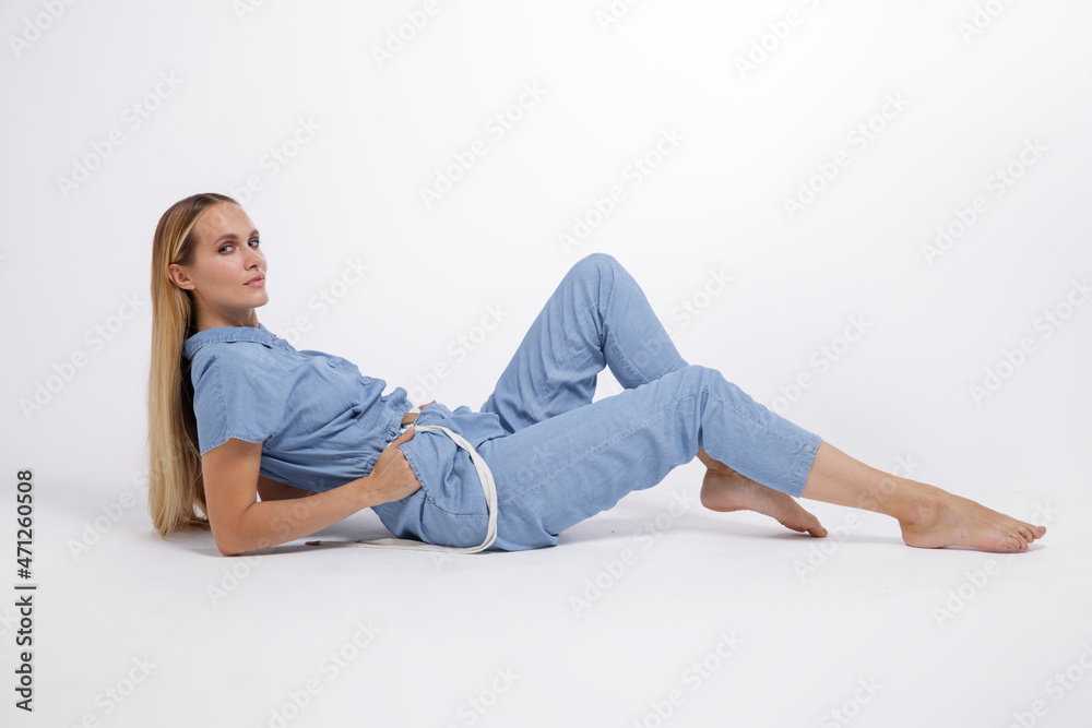 High fashion photo of a beautiful elegant young woman in a pretty blue jumpsuit, barefoot posing over white, soft gray background. Studio Shot, portrait. The model is sitting in the background. Blonde High fashion photo of a beautiful elegant young woman in a pretty blue jumpsuit, barefoot posing over white, soft gray background. Studio Shot, portrait. The model is sitting in the background. Blonde