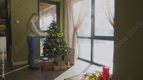 A beautiful young blonde girl in a sweater approaches the Christmas tree and puts on a Christmas ball. A Christmas tree by the window in the house with a bunch of gifts under it.