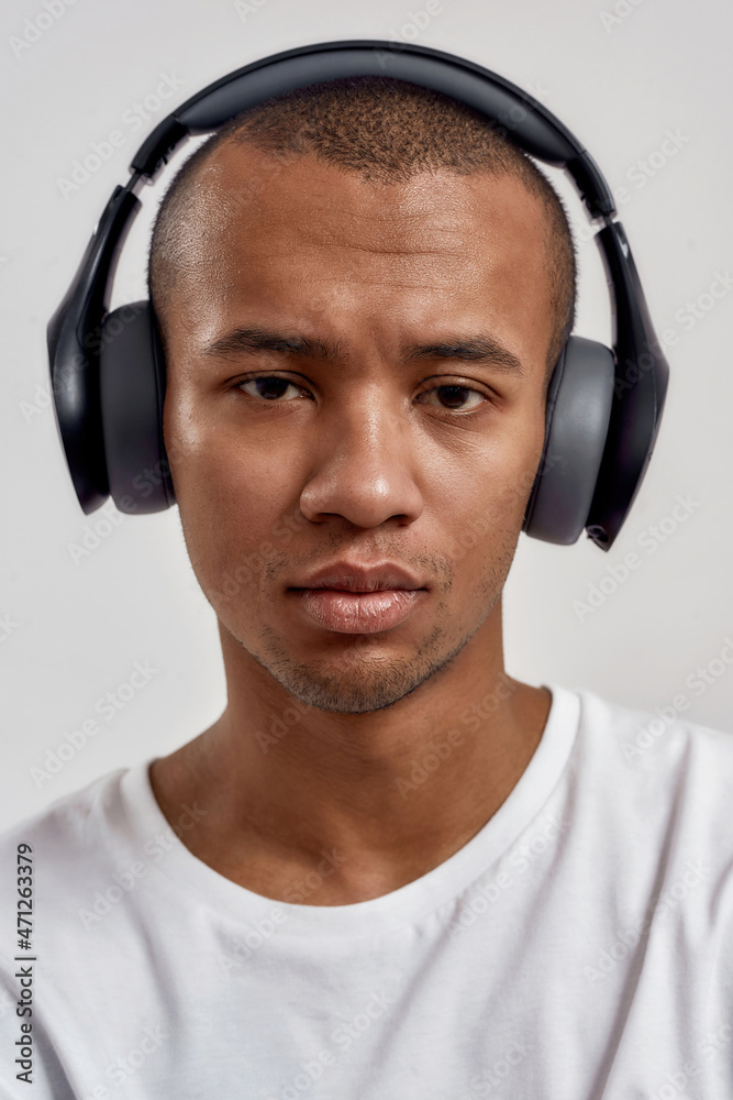 Closeup portrait of serious mixed race guy wearing headphones looking at camera, listening to music or podcast while posing isolated over white background Closeup portrait of serious mixed race guy wearing headphones looking at camera, listening to music or podcast while posing isolated over white background
