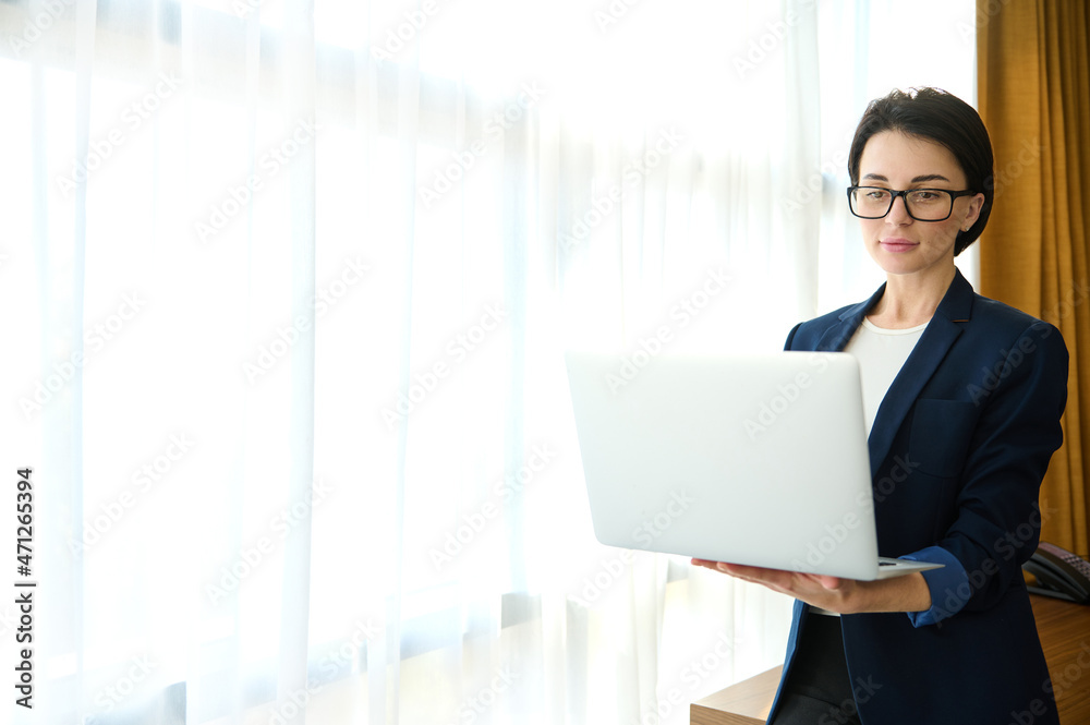 Prosperous concentrated woman, entrepreneur, journalist, sales manager in business casual suit working on laptop standing by window in hotel room. Cope space for advertisement text Prosperous concentrated woman, entrepreneur, journalist, sales manager in business casual suit working on laptop standing by window in hotel room. Cope space for advertisement text
