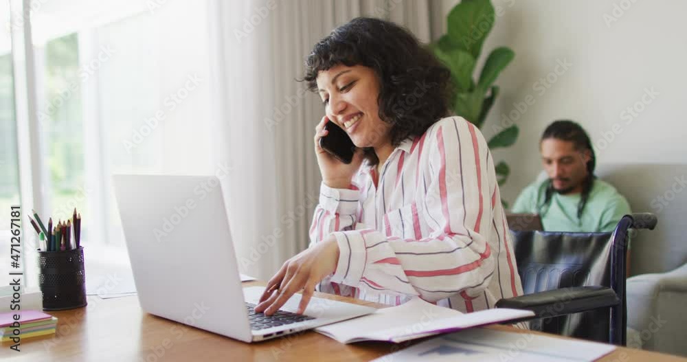 Happy biracial woman in wheelchair talking on phone using laptop, with male partner in living room Happy biracial woman in wheelchair talking on phone using laptop, with male partner in living room