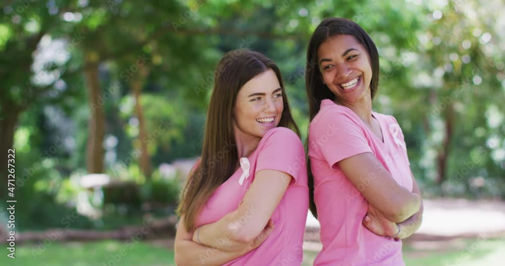 Portrait of two smiling diverse women in pink t shirts and cancer ribbons, back to back in park Portrait of two smiling diverse women in pink t shirts and cancer ribbons, back to back in park
