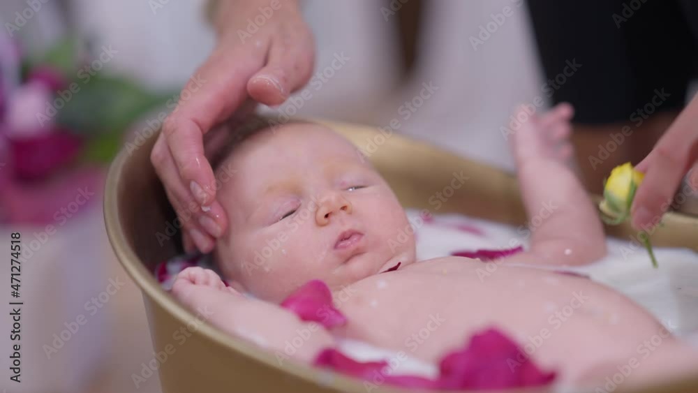 Charming little baby girl moving hands and legs lying in white milk water with mother hands putting yellow roses in bath with red petals. Portrait of adorable newborn Caucasian infant bathing indoors Charming little baby girl moving hands and legs lying in white milk water with mother hands putting yellow roses in bath with red petals. Portrait of adorable newborn Caucasian infant bathing indoors