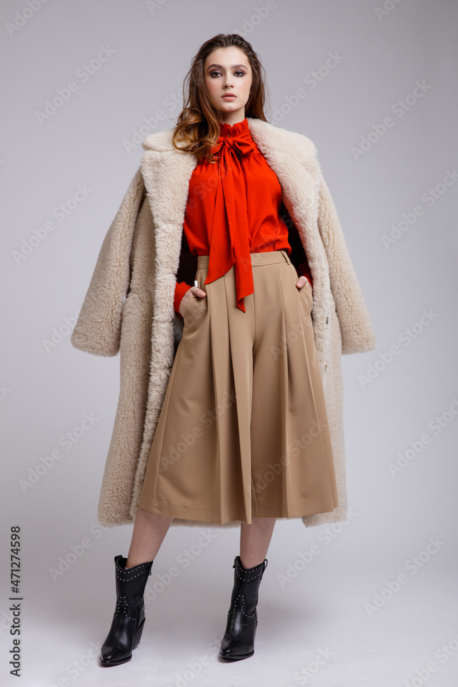 High fashion photo of a beautiful elegant young woman in a pretty beige coat, long skirt, red bow blouse, boots posing over white, soft gray background. Brown hair. Studio Shot, portrait. High fashion photo of a beautiful elegant young woman in a pretty beige coat, long skirt, red bow blouse, boots posing over white, soft gray background. Brown hair. Studio Shot, portrait.