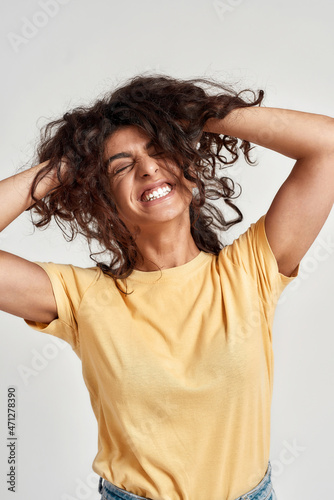 Portrait of excited young woman with dark curly hair in casual wear smiling with eyes closed at camera, adjusting her hair while standing isolated over gray background