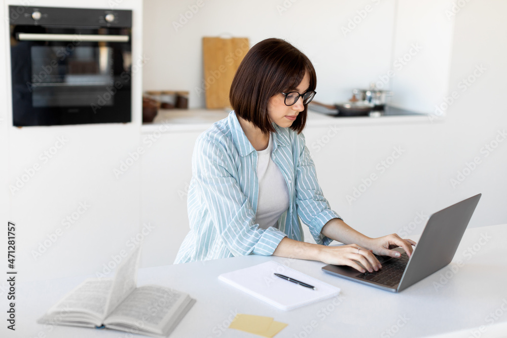 Remote business. Young freelancer lady working on laptop in kitchen, sitting at table and typing on pc keyboard Remote business. Young freelancer lady working on laptop in kitchen, sitting at table and typing on pc keyboard