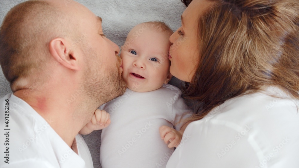 Happy family. Newborn baby with happy parents, top view. Healthy newborn baby in a white t-shirt with mom and dad. Close up Faces of the mother, father and infant baby. Cute Infant boy and parents Happy family. Newborn baby with happy parents, top view. Healthy newborn baby in a white t-shirt with mom and dad. Close up Faces of the mother, father and infant baby. Cute Infant boy and parents