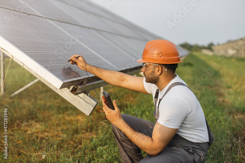 Competent technician in safety helmet and glasses measuring amperage of solar panels with multimeter. Indian man repairing photovoltaic cells outdoors. Green energy concept.