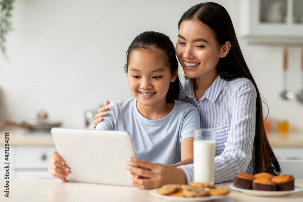 Happy asian mother and her daughter using digital tablet, looking for new recipes while sitting at kitchen, free space Happy asian mother and her daughter using digital tablet, looking for new recipes while sitting at kitchen, free space
