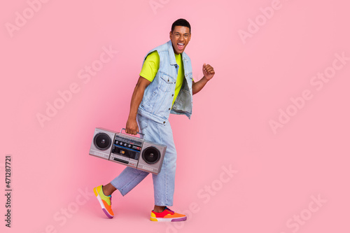 Full length profile photo of impressed millennial brunet guy go hold boom box wear t-shirt vest jeans sneakers isolated over pink color background