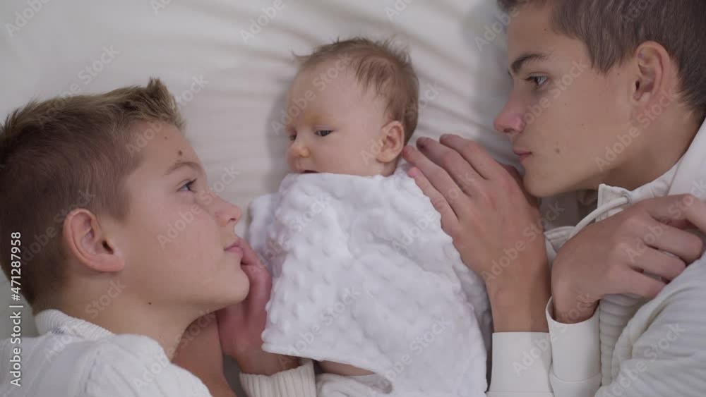 High angle view charming adorable infant girl lying on bed with teenage boys admiring child. Carefree Caucasian baby sister and brothers at home indoors. Slow motion High angle view charming adorable infant girl lying on bed with teenage boys admiring child. Carefree Caucasian baby sister and brothers at home indoors. Slow motion