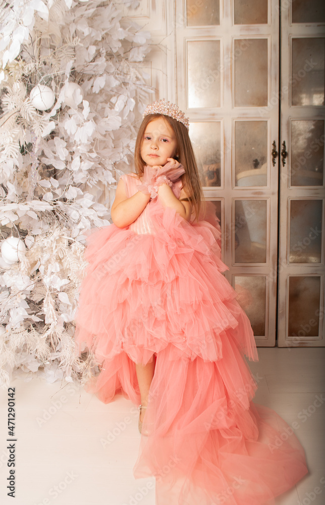 Fototapeta premium charming little girl stands against the background of snow-covered branches in a lush red dress for the New Year and Christmas 2022 near the mirror
