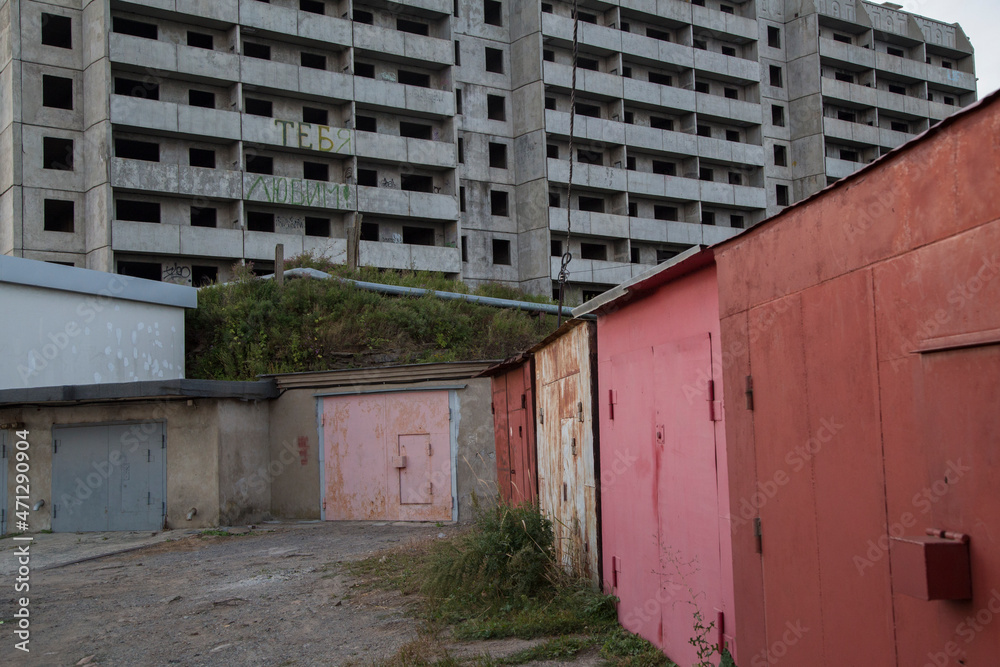 Fototapeta premium VLADIVOSTOK, RUSSIA. a soviet empty abandoned unfinished haunted residential concrete panel house building with balconies and a writing WE LOVE YOU in Russian and old red garages in front