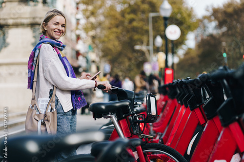 Woman taking a red bicycle in a bike rental station in the city with bike sharing app. Ecological way of urban commuting