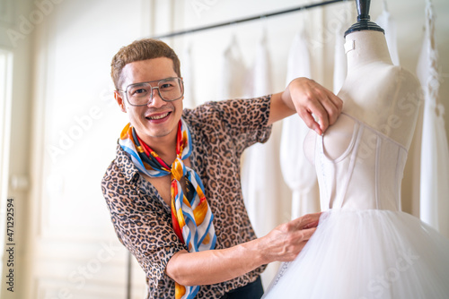 Fotografia, Obraz Portrait of Asian LGBTQ guy bridal shop owner using tape measure measuring wedding dress on sewing mannequin at wedding studio