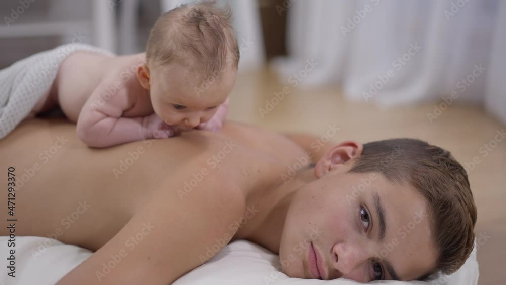 Confident Caucasian teenage boy looking at camera with little infant baby girl lying on his back. Portrait of handsome brother and cute adorable sister at home indoors. Slow motion Confident Caucasian teenage boy looking at camera with little infant baby girl lying on his back. Portrait of handsome brother and cute adorable sister at home indoors. Slow motion