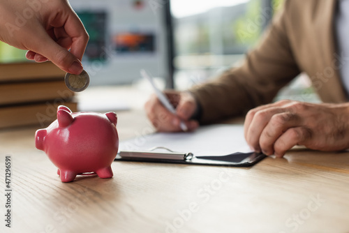 cropped view of woman holding coin near piggy bank while blurred businessman signing contract, anti-corruption concept